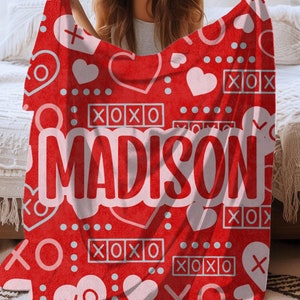 May include: Red fleece blanket with a white XOXO pattern and the name "MADISON" in large white letters. The blanket is folded in half and held by a person.