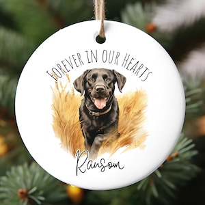 May include: A white, round Christmas ornament with a black Labrador Retriever illustration. The ornament has the text "FOREVER IN OUR HEARTS" and the name "Ransom" in script. The dog is set against a backdrop of tan-colored grass.