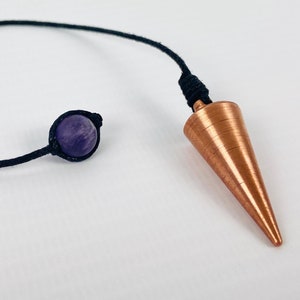 Puede incluir: Un péndulo de color cobre con forma cónica, conectado a un cordón negro. Una pequeña piedra redonda de amatista púrpura también está unida al cordón. El péndulo se utiliza para la adivinación y las prácticas espirituales.