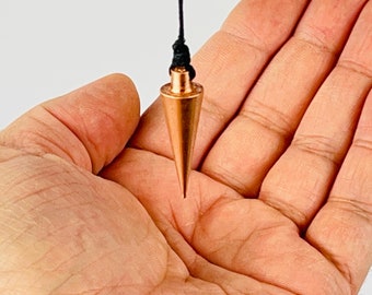 Könnte beinhalten: Ein kupferfarbenes Pendel mit konischer Form, das an einer schwarzen Schnur hängt. Das Pendel wird in einer Hand vor weißem Hintergrund gehalten. Das Pendel ist ein Werkzeug für Wahrsagerei und spirituelle Praktiken.