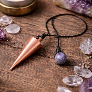 May include: A copper pendulum with a pointed tip and a black cord, next to a purple gemstone sphere. The image also features crystals, a lit candle, and amethyst stones, all arranged on a wooden surface.