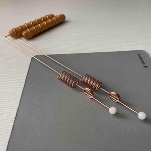 May include: Two copper dowsing rods with coiled centers and white stone tips, along with two wooden dowels. The rods are on a gray surface. The wooden dowels are light brown.