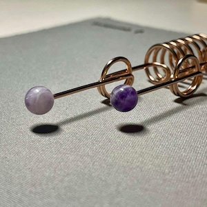 May include: Close-up of a copper-colored hair accessory with two gemstone beads. One bead is a light purple color, and the other is a darker purple. The accessory has a spiral design and is likely used to hold hair in place.