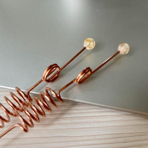 Può includere: Due forcine per capelli fatte a mano color rame. Ogni forcina presenta un design a spirale, un accento ad anello e una piccola gemma rotonda e chiara sulla parte superiore. Le forcine sono esposte su una superficie grigio chiaro.