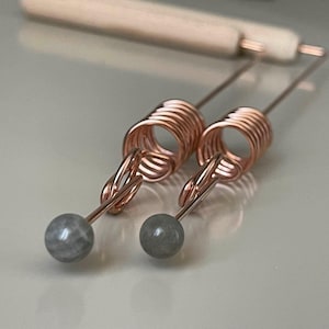 Könnte beinhalten: Ein Paar kupferfarbene Ohrringe mit einem spiralförmigen Design und einer grauen, kugelförmigen Perle am unteren Ende. Die Ohrringe haben ein zartes, handgefertigtes Aussehen, mit einem hellen Holzwerkzeug im Hintergrund.