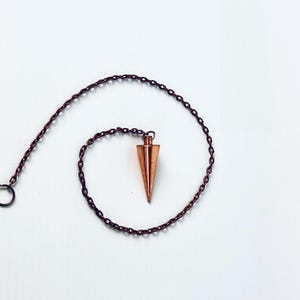 May include: A copper pendulum with a pointed tip hangs from a chain. The pendulum is a popular tool for divination and dowsing.