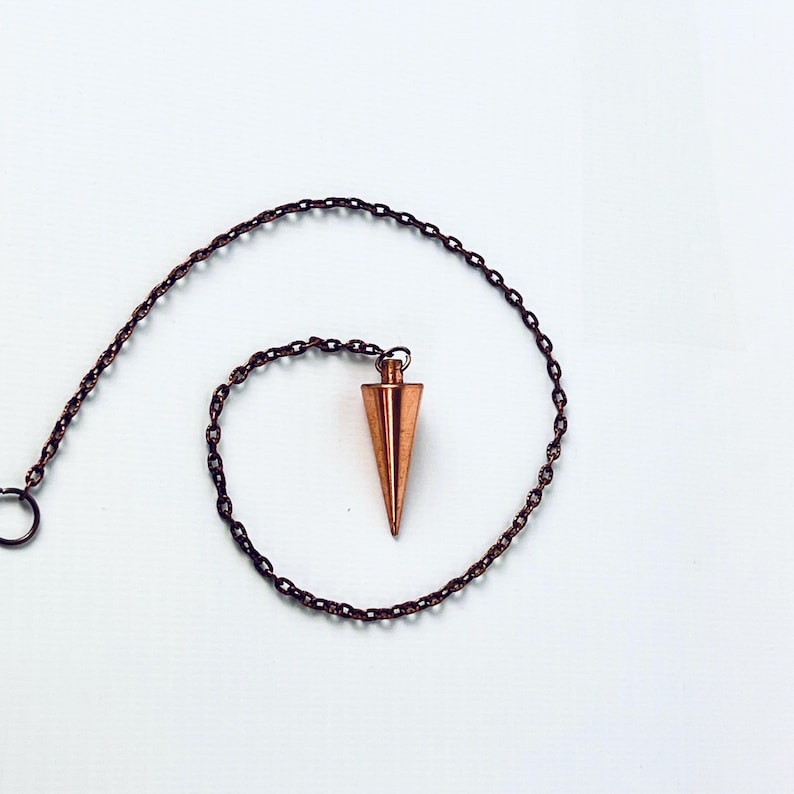 May include: A copper pendulum with a pointed tip hangs from a chain. The pendulum is a popular tool for divination and dowsing.