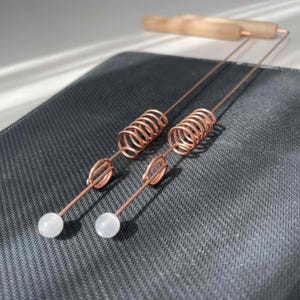 May include: Two copper dowsing rods with white crystal spheres at the ends. The rods have a spring and loop design, and a wooden handle. The rods are on a dark gray textured surface.