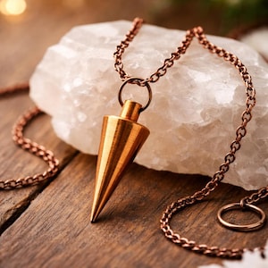 May include: A copper-colored pendulum necklace with a conical pendant. The pendant hangs from a matching chain, resting on a white, crystalline stone. The necklace is displayed on a wooden surface.