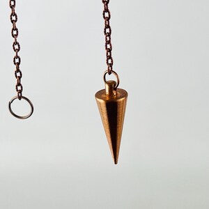 May include: A copper-colored pendulum with a pointed tip hangs from a chain. The chain is attached to a small ring.