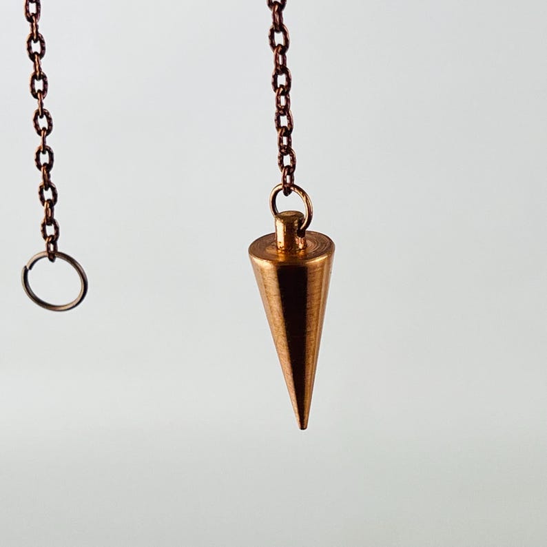 May include: A copper-colored pendulum with a pointed tip hangs from a chain. The chain is attached to a small ring.