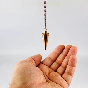 May include: A copper-colored pendulum with a pointed tip hangs from a chain. The pendulum is suspended above an open hand.