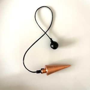 May include: A copper pendulum with a black weighted bob, attached to a black cord. The pendulum is pointed and has a shiny finish.