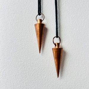 Pode incluir: Dois pêndulos de cobre pendurados em cordões pretos contra um fundo branco.