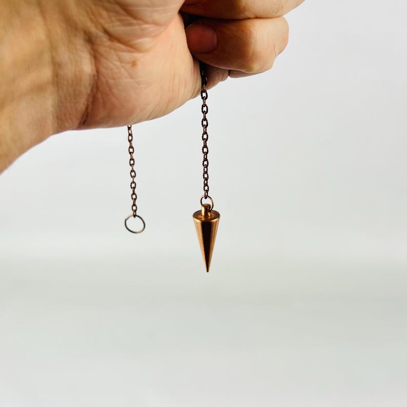 May include: A copper-colored pendulum with a pointed tip hangs from a chain. The chain has a small ring at the top.