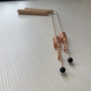 May include: A pair of copper-coloured earrings. Each earring features a spiral design and a small black bead. The earrings have a wooden bar at the top. They are displayed on a white surface.