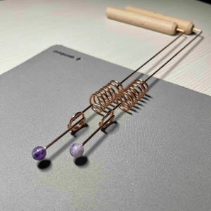 Puede incluir: Dos herramientas de alambre de color cobre con diseños en espiral y bucle, cada una con una pequeña gema redonda de color púrpura en el extremo. Dos clavijas de madera están en el fondo. Las herramientas están sobre una superficie gris.