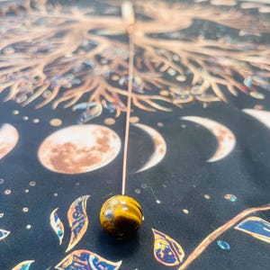 Puede incluir: Un péndulo de ojo de tigre dorado y marrón que cuelga de una fina cadena dorada sobre un fondo negro con un diseño de árbol de la vida dorado y marrón.