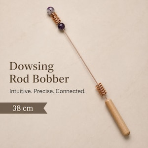 May include: A dowsing rod bobber, 38 cm long, with a wooden handle and a copper wire shaft. The rod features a copper coil and two purple and white gemstone beads. Text on the image reads "Dowsing Rod Bobber. Intuitive. Precise. Connected."