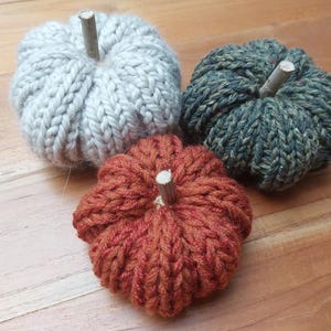 Op de afbeelding: Drie handgebreide pompoenen in grijstinten, oranje en groen, elk met een kleine houten steel. De pompoenen zijn geplaatst op een houten oppervlak, perfect voor herfstdecoratie.