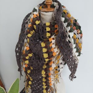 Op de afbeelding: Een handgemaakte gehaakte sjaal in een driehoekige vorm, weergegeven op een mannequin. De sjaal heeft een patchwork-ontwerp met vierkanten in tinten bruin, geel, oranje, groen en wit. De randen zijn afgewerkt met franjes.