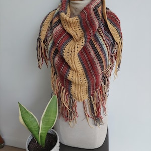 Op de afbeelding: Handgebreide sjaal in meerdere kleuren met franjes. De sjaal heeft rode, gouden en blauwe strepen. De sjaal wordt op een mannequin gepresenteerd. Een kleine potplant met groen-witte bladeren staat op de voorgrond.