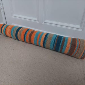 Pode incluir: Um batente de porta longo e listrado feito de tecido de malha. O batente de porta é laranja, marrom, azul-petróleo e bege. Está sobre um tapete bege.