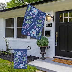 May include: A white house with a black door and a blue floral flag hanging on the front porch. The house number is 805. There are two potted plants on the porch.
