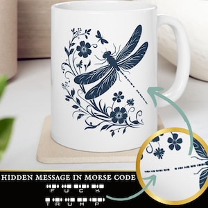 May include: White ceramic mug featuring a blue dragonfly and floral design. The dragonfly's wings contain a hidden message in Morse code that reads "F U C K T R U M P".