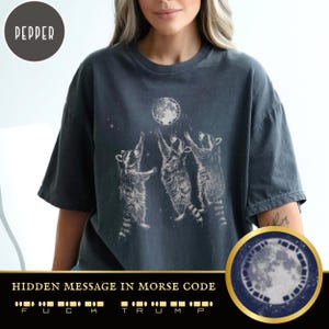 May include: A dark gray t-shirt with a graphic of three raccoons reaching up to a full moon. The moon has a hidden message in Morse code that reads "F U C K T R U M P".