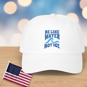 May include: A white baseball cap with the text "BE LIKE WATER NOT ICE" in blue. The cap is on a wooden surface with a small American flag in the foreground. The background has a bokeh effect.
