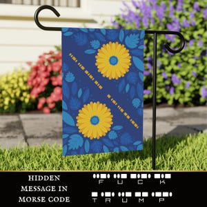 Könnte beinhalten: Eine blaue Gartenflagge mit einem Blumenmuster mit gelben Blumen und grünen Blättern. Die Flagge hat eine versteckte Botschaft in Morsecode, die "F.U.C.K. TRUMP" lautet.