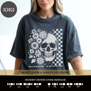 May include: Dark gray t-shirt with a skull and floral design. The graphic includes a skull, daisies, and a checkered pattern. The text "SELECT FROM A VARIETY OF COLORS!" is displayed. The hidden Morse code message reads "FUCK TRUMP".