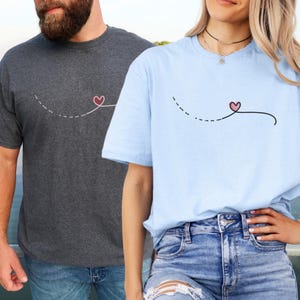 Puede incluir: Dos camisetas con un diseño simple de un corazón conectado por una línea punteada. La camiseta de la izquierda es gris oscuro y la camiseta de la derecha es azul claro.