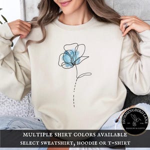 May include: A beige sweatshirt with a blue watercolor flower design. The text "Hummingbird Lane" is on the bottom right corner of the sweatshirt. The text "Multiple Shirt Colors Available Select Sweatshirt, Hoodie or T-Shirt" is at the bottom of the image.