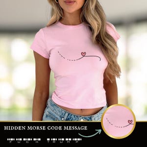 May include: Pink t-shirt with a small heart and a dashed line design. The shirt has a hidden Morse code message that reads "F.U.C.K. TRUMP".