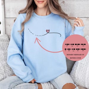 May include: Light blue sweatshirt with a black line design featuring a heart and arrow. The sweatshirt has a hidden message in Morse code that reads "FUCK TRUMP". The text is displayed on a pink circle.