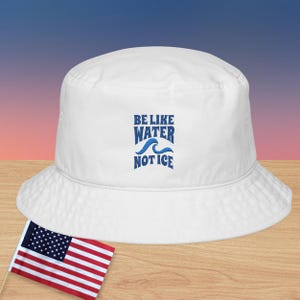May include: White bucket hat with the text "BE LIKE WATER NOT ICE" in blue. The hat is made of a soft material and has a wide brim. An American flag is in the foreground.