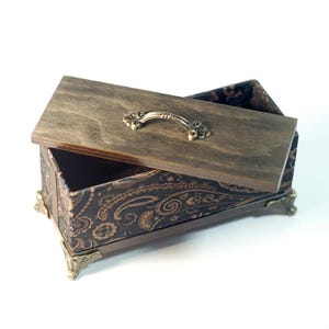 Puede incluir: Una caja de madera con tapa con bisagras, con un acabado marrón oscuro y un patrón de cachemira decorativo en el interior. La tapa tiene un asa de metal y la caja tiene patas decorativas de metal.
