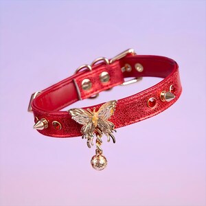 May include: A red leather choker with gold studs and a dangling gold butterfly charm. The butterfly has a yellow center and is attached to a small gold bell.