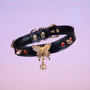 May include: Black leather choker with gold accents, featuring a dangling butterfly charm with red gemstones and a small ball at the end.