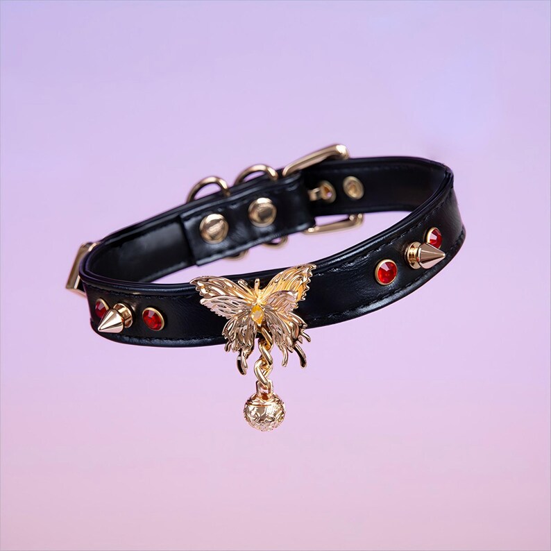 May include: Black leather choker with gold accents, featuring a dangling butterfly charm with red gemstones and a small ball at the end.