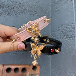 May include: Two dog collars with butterfly charms. The top collar is pink with pink rhinestones and the bottom collar is black with red rhinestones.