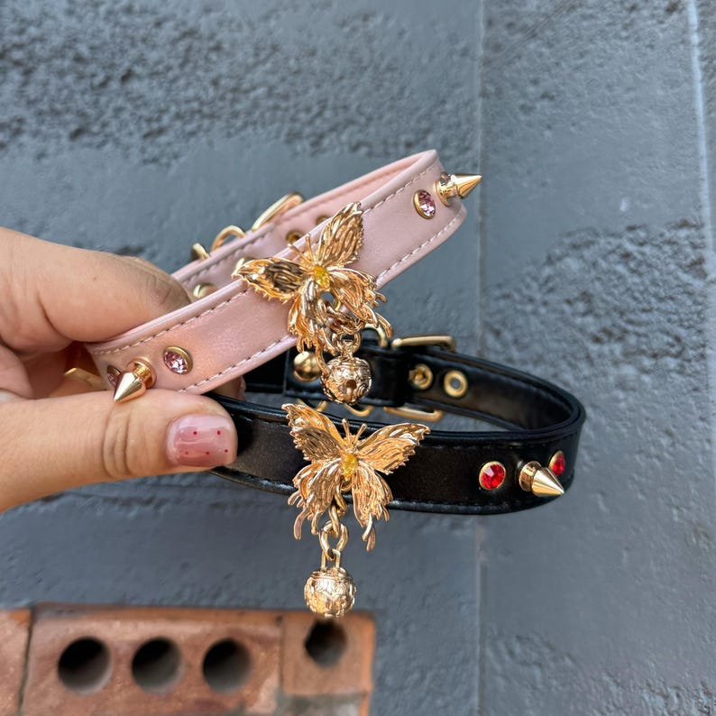 May include: Two dog collars with butterfly charms. The top collar is pink with pink rhinestones and the bottom collar is black with red rhinestones.