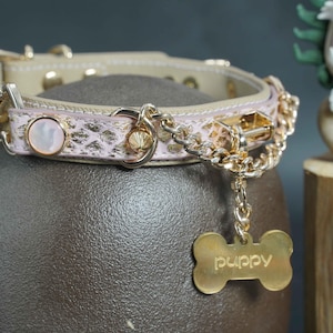 Puede incluir: Un collar de perro de cuero rosa con una cadena dorada y una etiqueta en forma de hueso que dice "puppy".