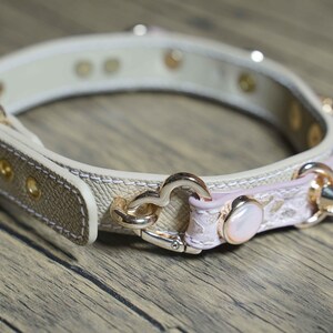 Puede incluir: Un collar de perro de cuero beige con detalles en rosa y herrajes dorados. El collar tiene un dije en forma de coraz&oacute;n y un peque&ntilde;o dije de perla.