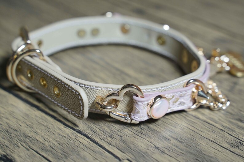Puede incluir: Un collar de perro de cuero beige con detalles en rosa y herrajes dorados. El collar tiene un dije en forma de coraz&oacute;n y un peque&ntilde;o dije de perla.