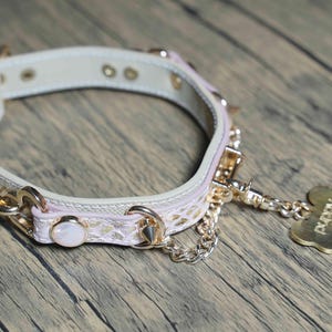 Puede incluir: Un collar de perro de cuero blanco con detalles en rosa y herrajes dorados. El collar tiene una cadena unida a una etiqueta dorada en forma de hueso que dice "Puppy".