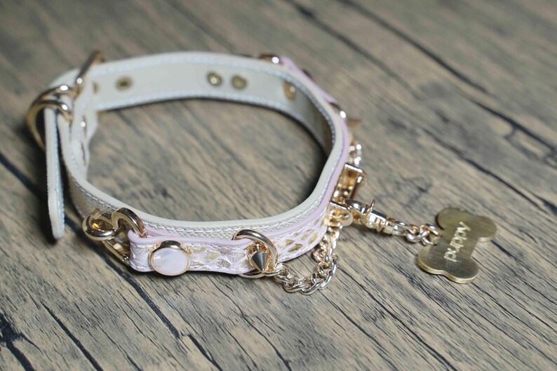 Puede incluir: Un collar de perro de cuero blanco con detalles en rosa y herrajes dorados. El collar tiene una cadena unida a una etiqueta dorada en forma de hueso que dice "Puppy".