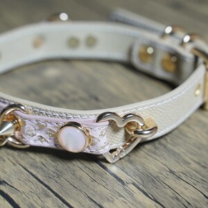 Puede incluir: Un collar gargantilla de cuero blanco con ribete de encaje rosa y detalles dorados. El collar presenta un dije en forma de coraz&oacute;n, un bot&oacute;n perlado y un detalle de punta.